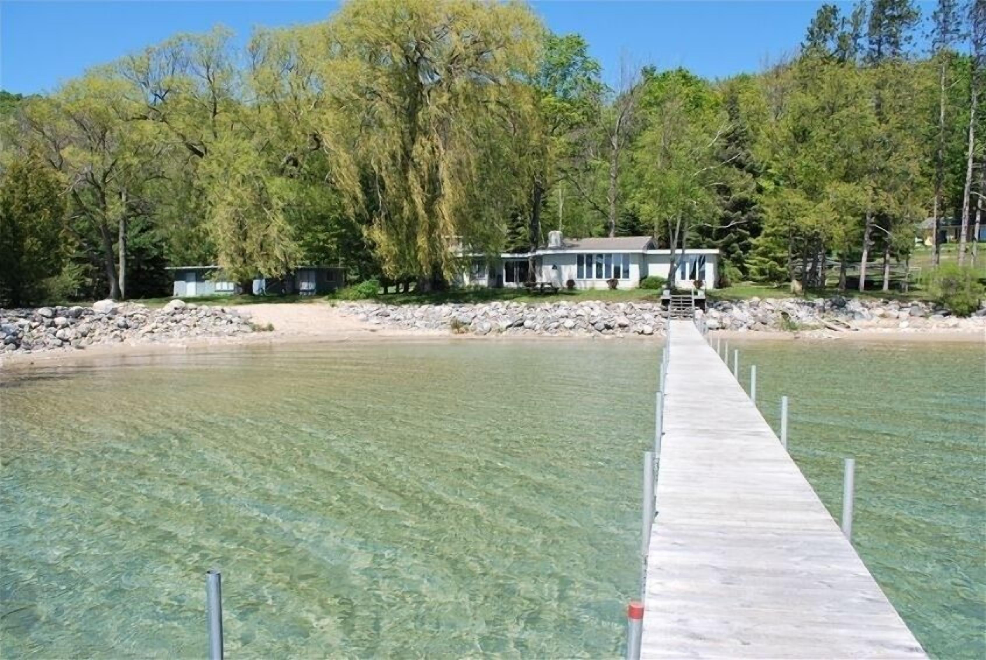Un muelle de madera que se adentra en un lago transparente, con una casa y árboles al fondo.