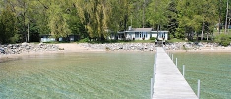 Un muelle de madera que se adentra en un lago transparente, con una casa y árboles al fondo.