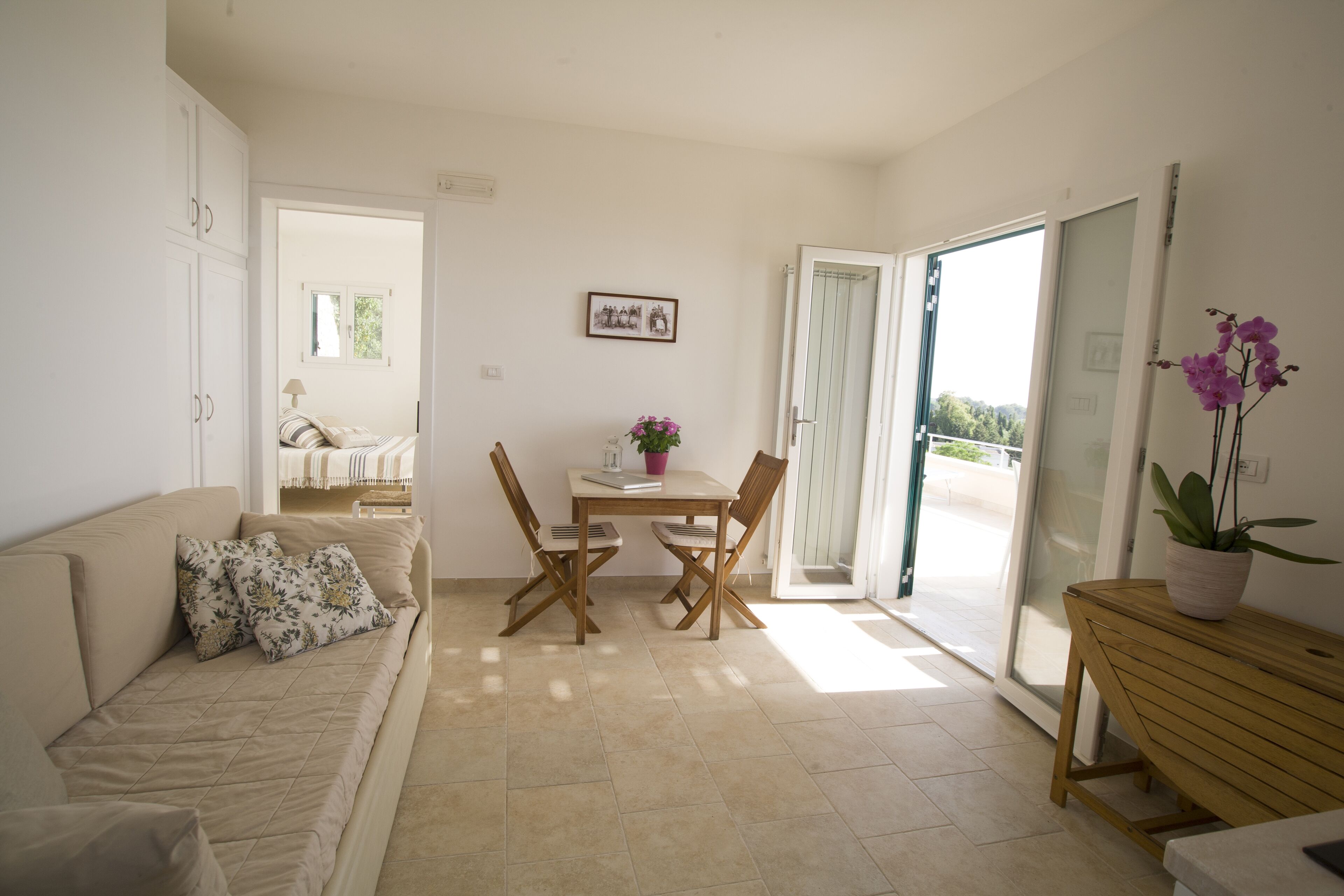 A living room with a sofa, a dining table with chairs, a small table with a potted plant, and a sliding glass door leading to a balcony.