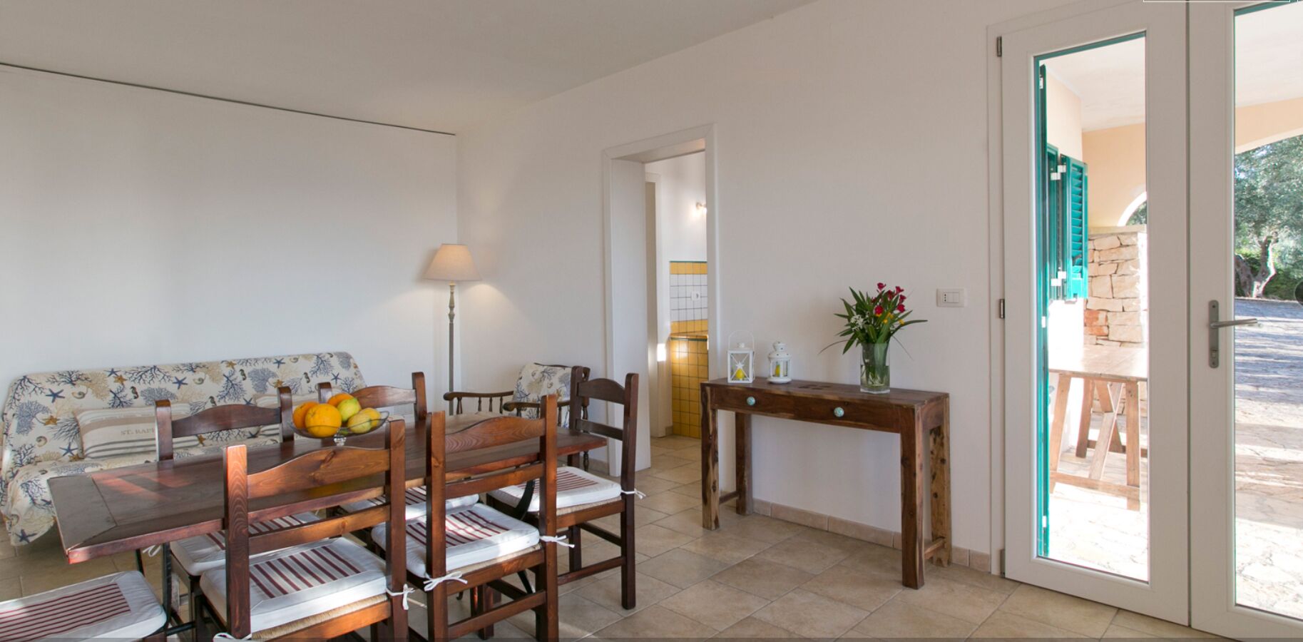 Uma área de refeições com uma mesa e cadeiras de madeira, um sofá com padrão floral, um abajur e um vaso com flores.