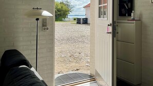Una habitación con pared de ladrillo blanco, un sofá negro, una lámpara de pie y una puerta que da a un área de grava con vistas al mar.