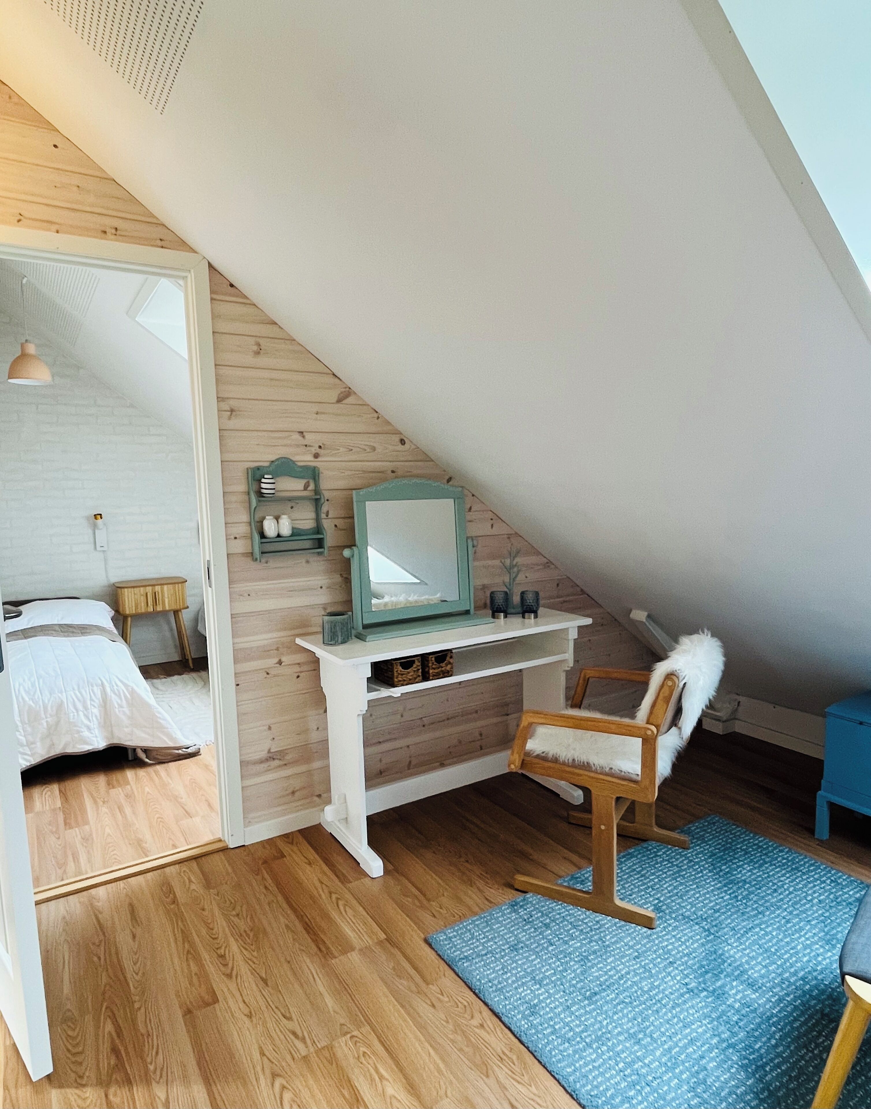 Un dormitorio acogedor en un ático con techo inclinado, paredes de madera, una cama blanca, un escritorio con espejo y una alfombra azul.