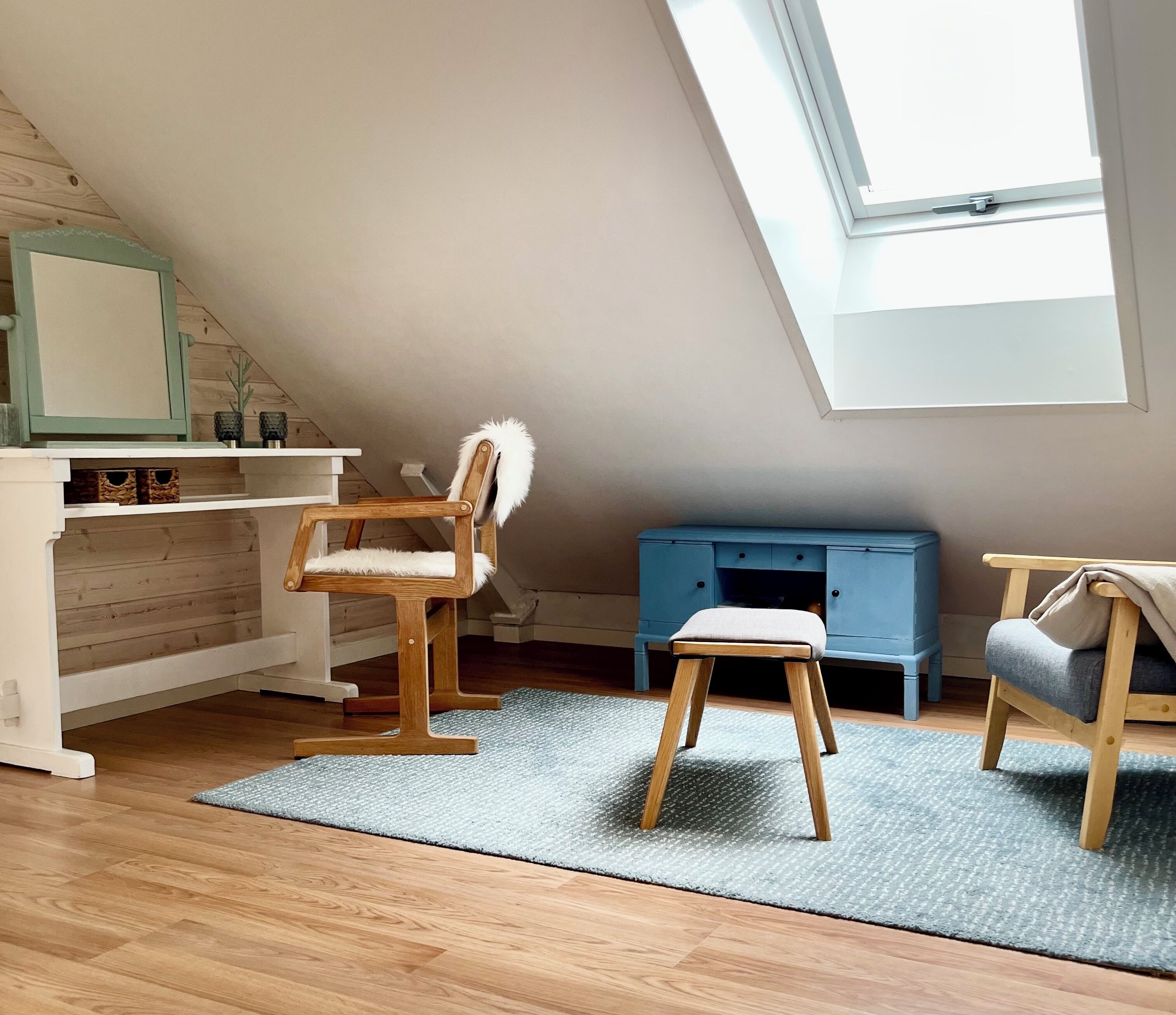 Une chambre à coucher aménagée dans un grenier, avec un plafond en pente, un plancher en bois, un puits de lumière, un bureau, une chaise, un petit tabouret et une armoire bleue.
