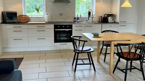 Una cocina moderna con armarios blancos, un comedor con mesa y sillas de madera, y dos ventanas grandes con vistas a follaje.