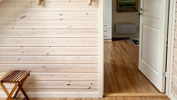 Una habitación con techo inclinado, paredes de madera, un estante pequeño con un adorno, un espejo de mimbre y un banco de madera.