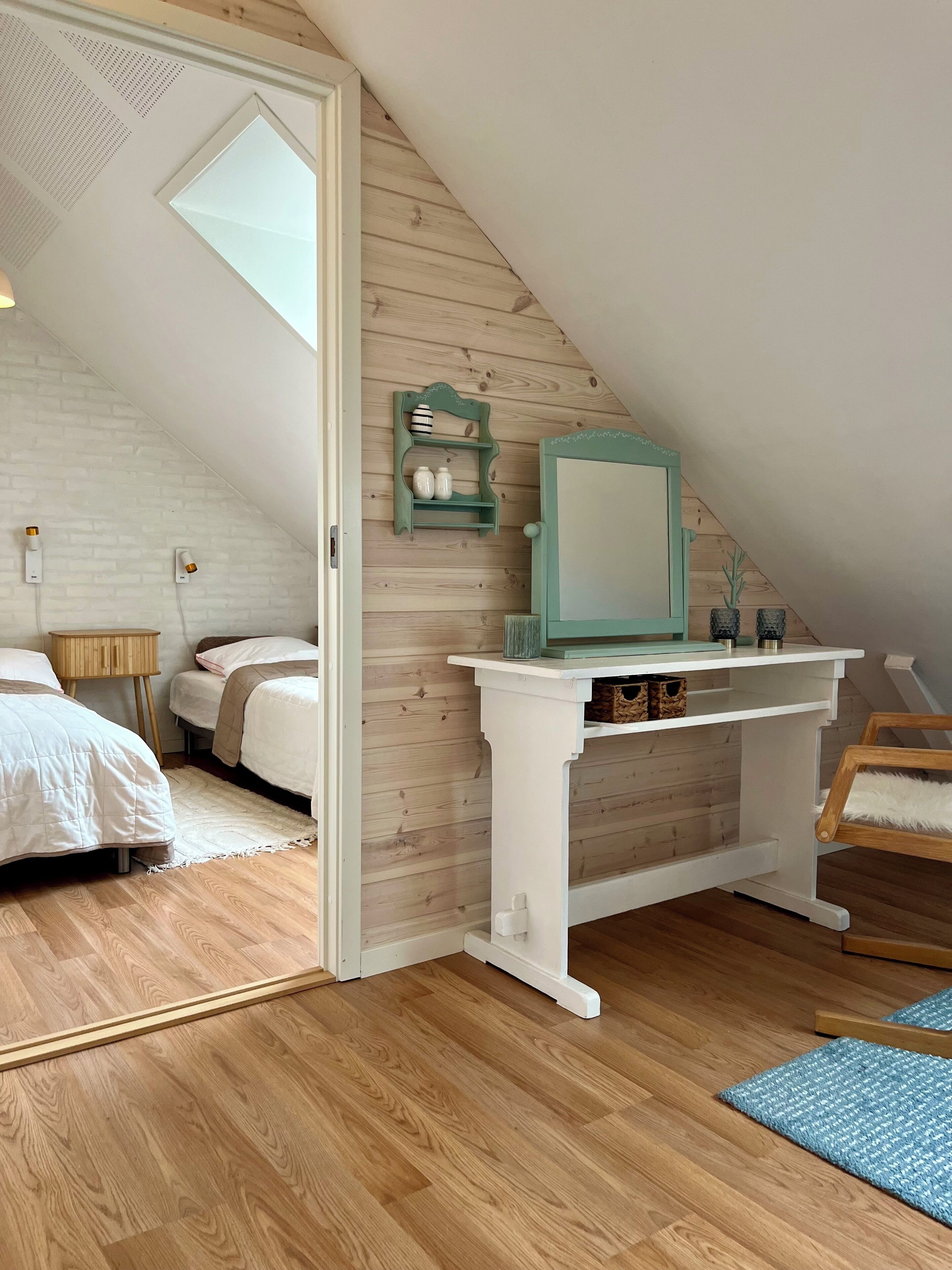 Une chambre avec un plafond en pente, un plancher en bois, un lit blanc, un petit bureau avec un miroir et une étagère ornée d’objets décoratifs.