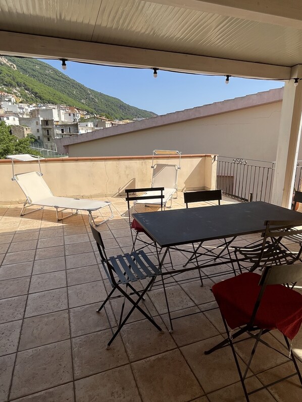 An outdoor terrace with a table and chairs, a view of a hillside town, and a covered area with a ceiling.