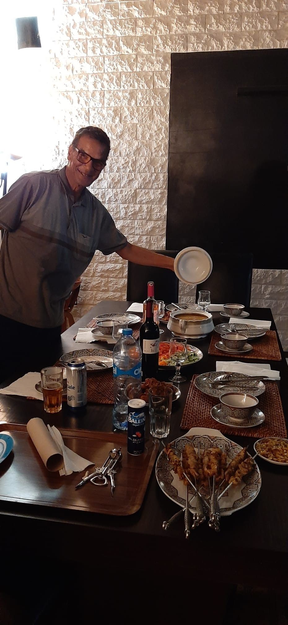 A dining table set with various dishes, utensils, and beverages, indicating a meal setup in a room with textured walls and a dark partition.