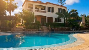 A two-story building with a swimming pool, surrounded by palm trees and a clear sky.