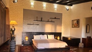 A bedroom with a large bed, a stone wall, a wooden ceiling, and a nightstand with a lamp.