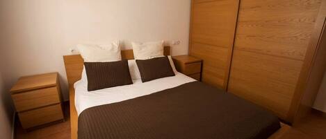A bedroom with a bed, two nightstands, a wooden wardrobe, and a white wall.