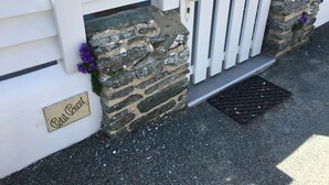 Un mur de pierre avec une porte blanche, une petite marche menant à la porte, et une enseigne sur laquelle on peut lire « Petit Cœur ».