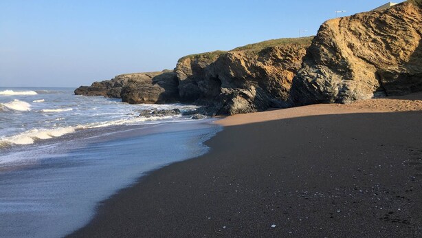 Une plage au sable sombre, avec des vagues et des falaises rocheuses.