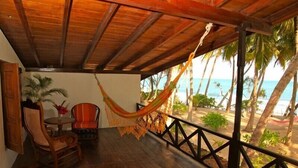 Um deck de madeira com uma rede, cadeiras e uma mesa, com vista para uma praia com palmeiras e o oceano.