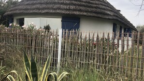 A thatched-roof house with a white exterior and a blue door, surrounded by a wooden fence and tropical vegetation.
