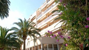 Um hotel com vários andares, varandas, janelas amplas e vegetação exuberante, incluindo palmeiras e plantas floridas.