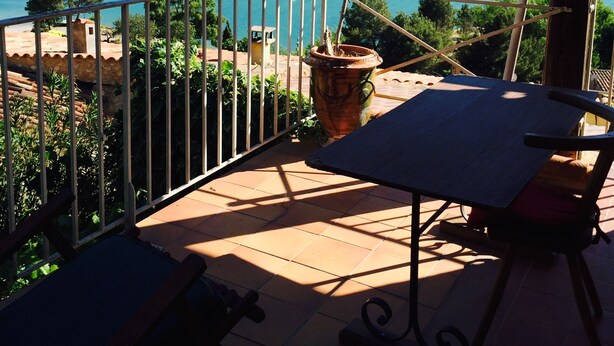 Ein Balkon mit Tisch und Stühlen mit Blick auf ein türkisblaues Meer und grüne Hügel.