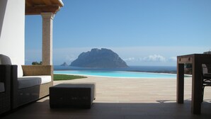 A patio with wicker furniture, a stone table, and a view of a clear blue sea and a distant island.