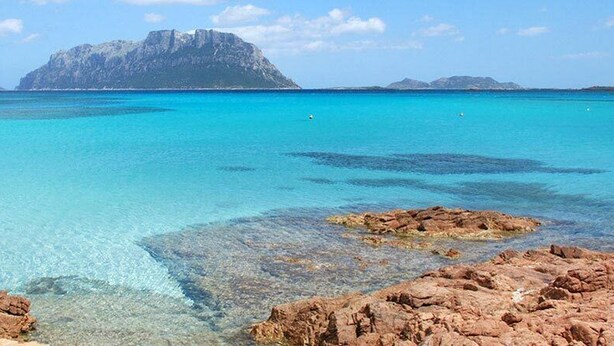 A coastal landscape with clear turquoise waters, rocky shorelines, and a prominent mountain in the background.