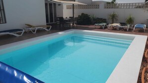 Piscina retangular com borda branca, rodeada por espreguiçadeiras e um espaço de estar com mesa e cadeiras.