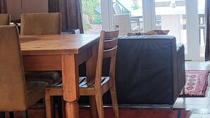 Uma área de refeições com mesa e cadeiras de madeira, um sofá e vista para um espaço exterior com plantas através de uma porta de vidro.
