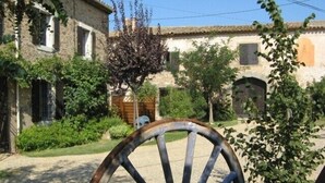 Un patio rústico con edificios de piedra, una gran rueda de metal y abundante vegetación.