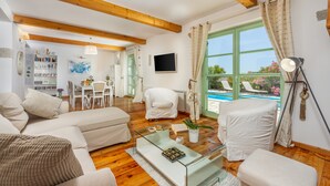 A modern living room with a wooden ceiling, a glass coffee table, white sofas, a dining area, and a view of a swimming pool.