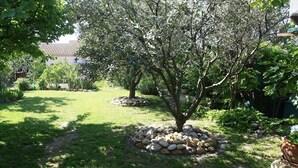 A garden with a tree surrounded by a circle of rocks, a grassy lawn, and a house in the background. - 200 meters from the heart of the village, App 55 M² WiFi In a closed courtyard, quiet (Châteauneuf-De-Gadagne)