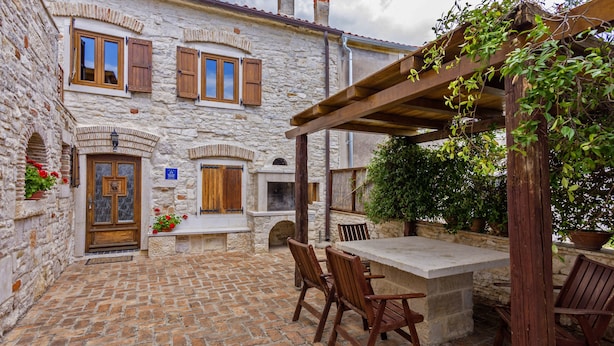 Un edificio de piedra con contraventanas de madera, un banco de piedra y una mesa con sillas en un patio al aire libre.