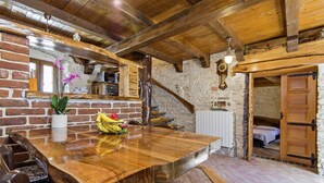 Une table de salle à manger rustique en bois, sur laquelle on trouve un panier de fruits, un vase avec des fleurs et un miroir au-dessus. On aperçoit un mur en pierre et une porte en bois à l’arrière-plan.