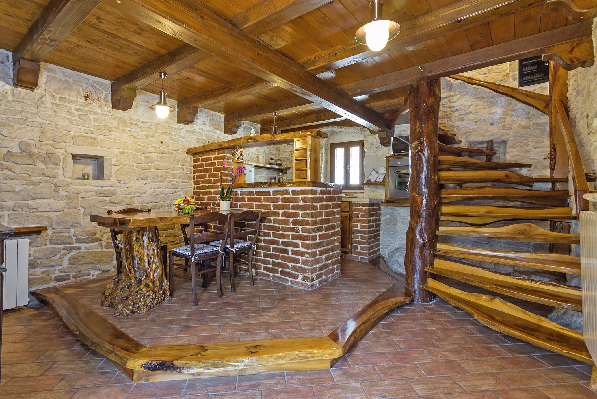 A rustic interior with a stone wall, wooden ceiling beams, a brick fireplace, and a wooden staircase.
