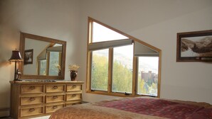 Un dormitorio con una cama grande, un aparador con cajones, un espejo, una ventana con vistas a árboles y edificios, y un cuadro enmarcado en la pared.
