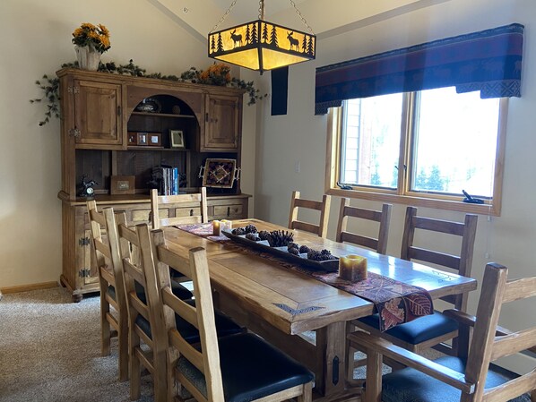 A dining area with a wooden table and chairs, a chandelier, and a window with a view of trees.