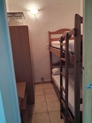 A small, compact room with a bunk bed, a wooden wardrobe, and a light fixture on the wall.