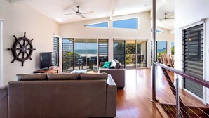 Amplia sala de estar con un sofá grande, televisión y una decoración con timón de barco. Hay puertas de cristal corredizas que dan a un balcón con vista al océano.