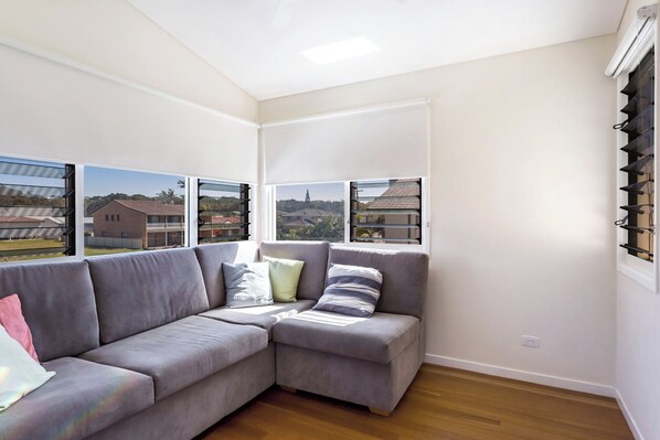 A modern living room with a grey sectional sofa, a small table, and a view of a residential area through large windows. - Tidemark backs on to the beautiful Diamond Beach, Ocean views at their finest (Diamond Beach)