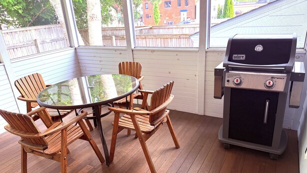 A wooden deck with a glass table and chairs, a Weber Spirit grill, and a view of a brick building through the windows. - Downtown Kennebunk Home  (Kennebunk)