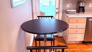 A compact kitchen with a dining area, including a round table and chairs, a coffee maker, and a view through a door to an outdoor deck. - Downtown Kennebunk Home  (Kennebunk)