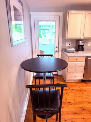 A compact kitchen with a dining area, including a round table and chairs, a coffee maker, and a view through a door to an outdoor deck. - Downtown Kennebunk Home  (Kennebunk)