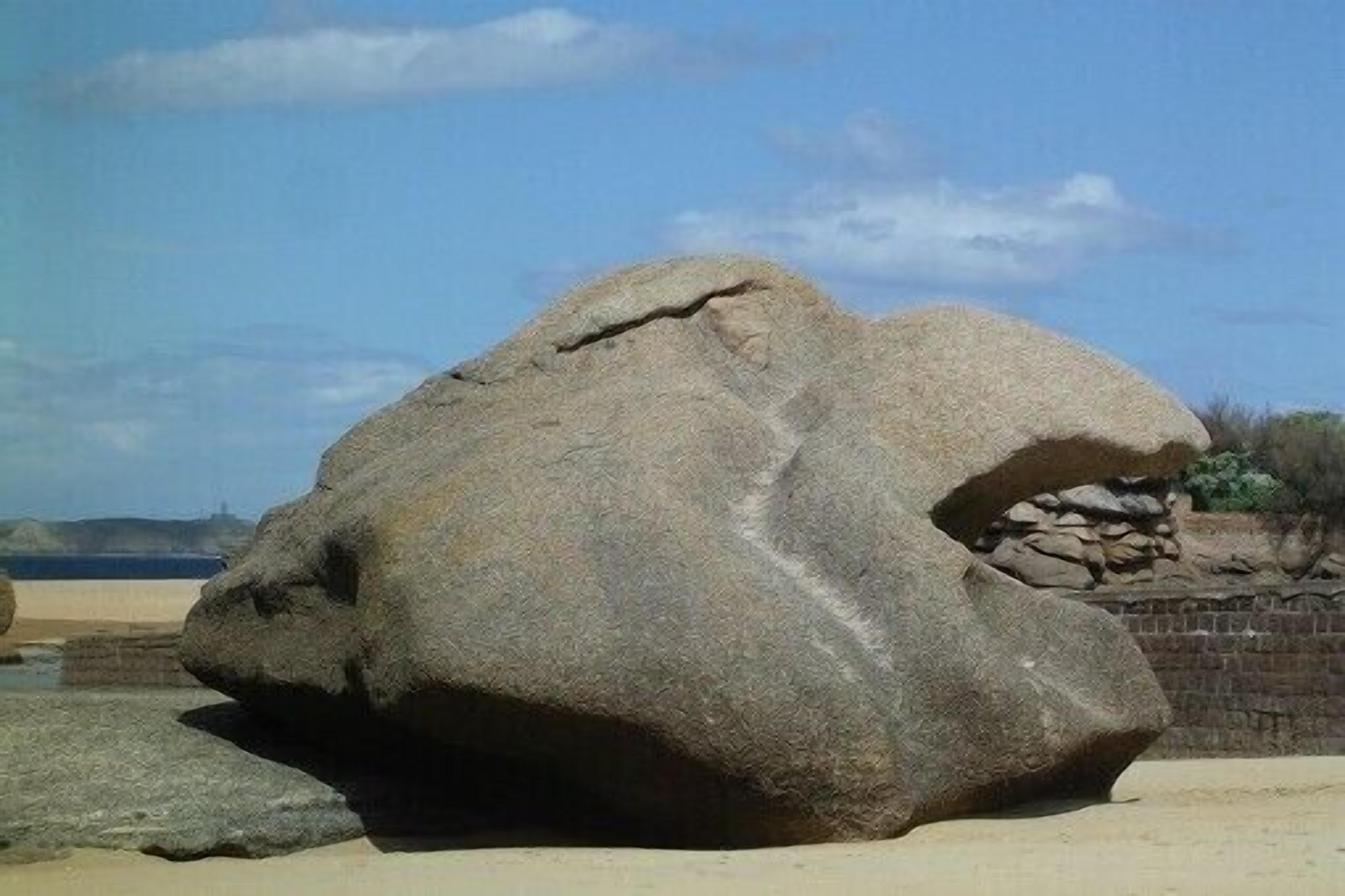 A large rock formation with a cave-like opening, situated on a sandy beach with a clear sky and distant hills.