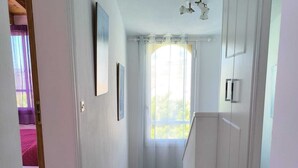 En smal gang med flisegulv, hvide vægge og et vindue med let gardin.