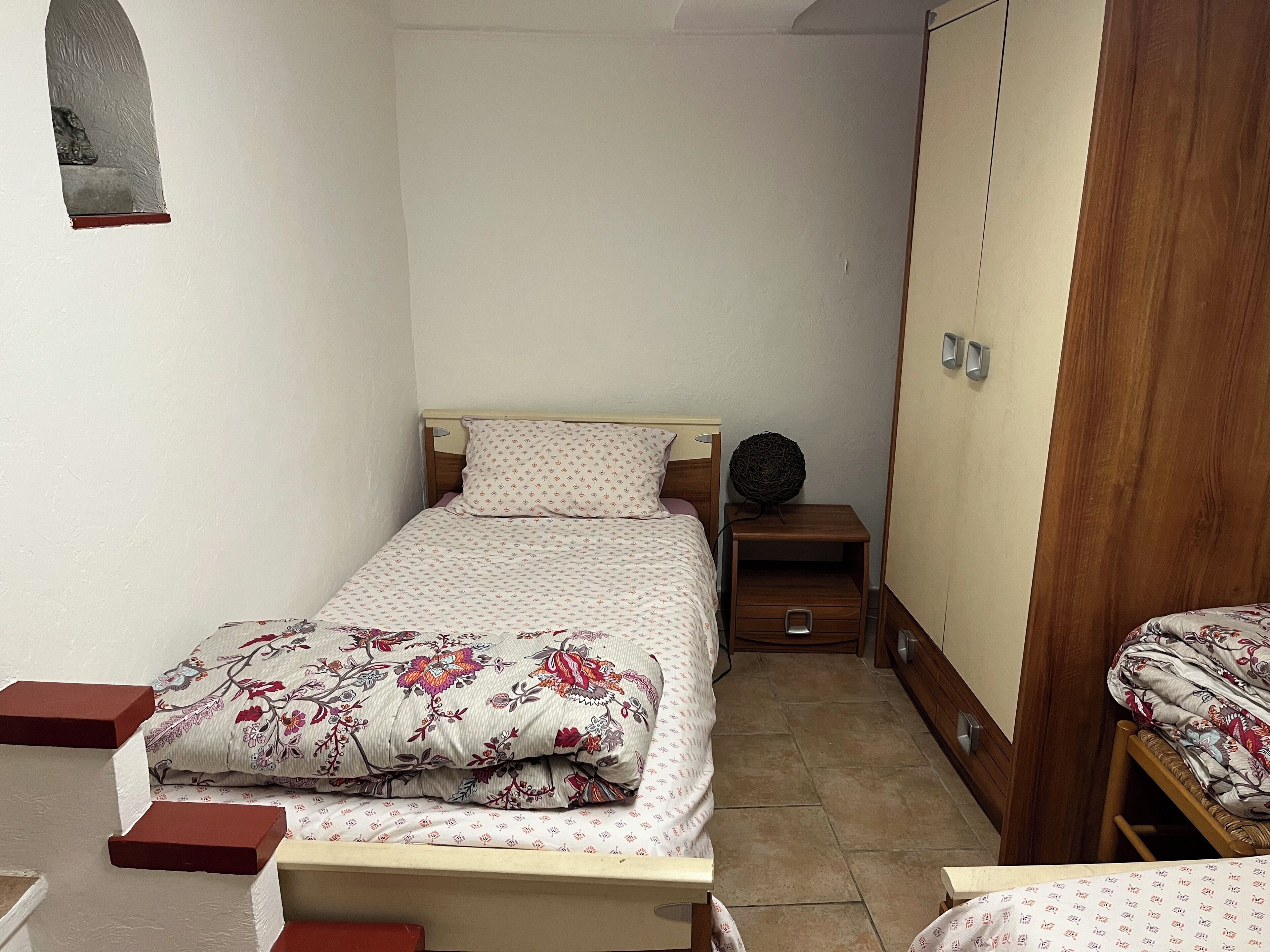 A small, neatly arranged bedroom with a single bed, a nightstand, a wardrobe, and a small window.