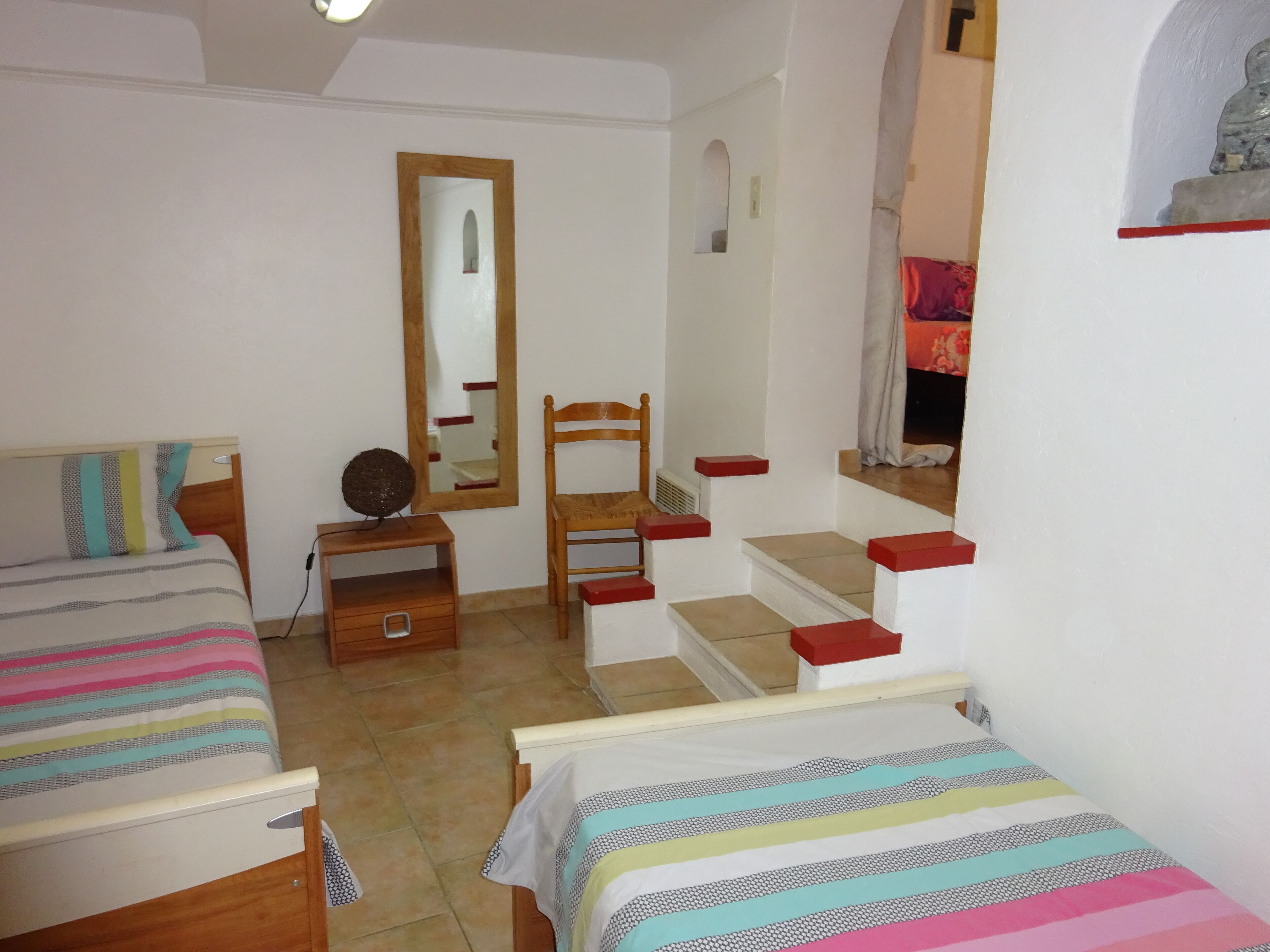 Une chambre avec deux lits, une chaise en bois, une petite table, un miroir et un escalier.