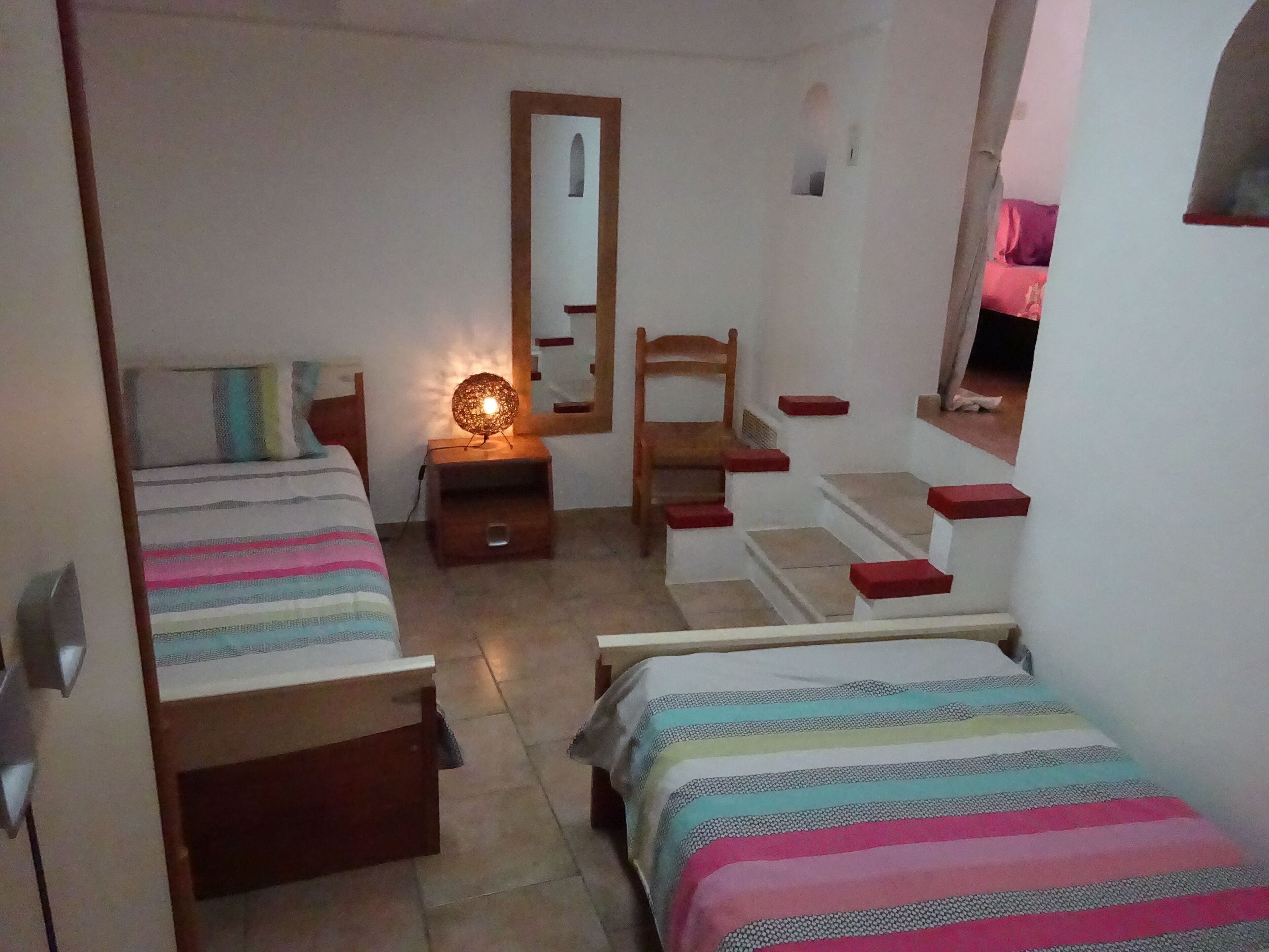 A bedroom with two beds, a wooden chair, a nightstand with a lamp, a mirror, and a window.
