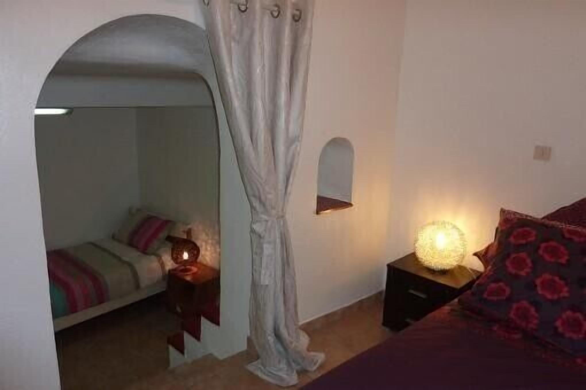 A bedroom with a bed, a nightstand with a lamp, a mirror, and a door leading to another room.
