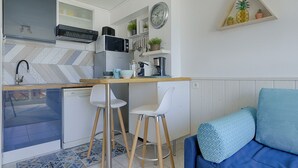 A compact kitchen with a microwave, kettle, and coffee maker. There is a small dining area with two stools and a blue sofa with patterned cushions.