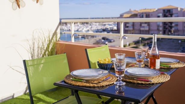 A table set for two with plates, glasses, and a bottle on a balcony overlooking a marina.