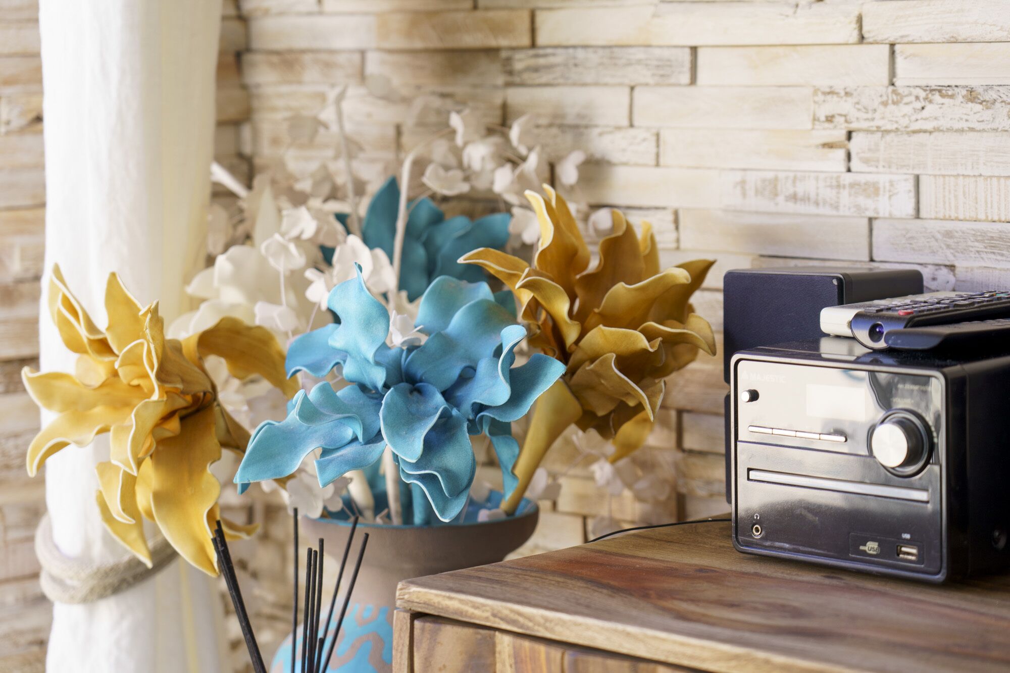 A wooden table with a radio, a bowl of artificial flowers, and a vase with a white column in the background.