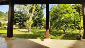 A covered outdoor area with a tiled floor, surrounded by trees and grass. - House for rent in gated community, Boiçucanga, São Sebastião, SP (São Sebastião)