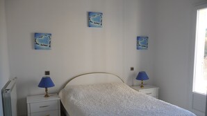 A bedroom with a bed, two bedside tables with lamps, a radiator, and three wall-mounted pictures. - Furnished T2 (3 stars) with terrace and pool in the Luberon Côté Sud. (La Tour-D'aigues)